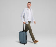 Male talent mid-stride, holding the trolley handle of The Front Pocket Carry-On in Coast Blue