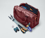 70L duffle bag packed with outdoor travel gear, including packing cubes, clothing, hiking poles, and binoculars. A water bottle and carabiner are attached to the exterior webbing loop.