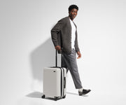 A 6'1" male model is standing next to and walking with The Bigger Carry-On suitcase by holding the raised handle to show size and scale