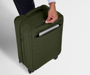 A person is unzipping the hidden exterior pocket on The Softside Carry-On in Olive Green, and there is a laptop peeking out of the second, deeper no-show pocket on the outside of the suitcase.