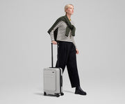 Female talent mid-stride, holding the trolley handle of The Front Pocket Carry-On in Cloud Gray