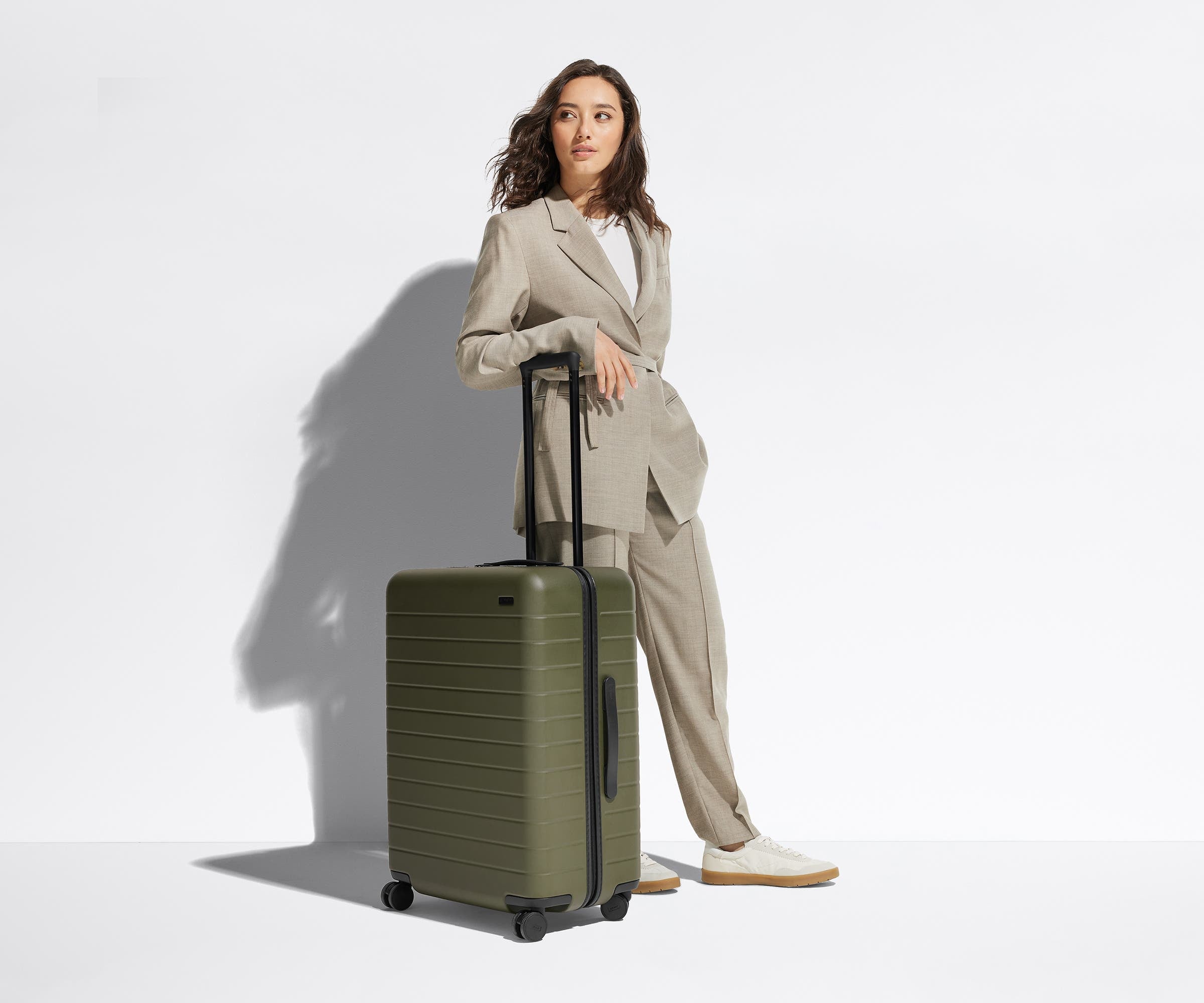 A 5'6" female model is standing next to The Medium suitcase and leaning her arm against the raised handle to show size and scale