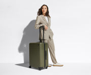 A 5'6" female model is standing next to The Medium suitcase and leaning her arm against the raised handle to show size and scale
