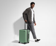 A 6'1" male model is standing next to and walking with The Bigger Carry-On suitcase by holding the raised handle to show size and scale