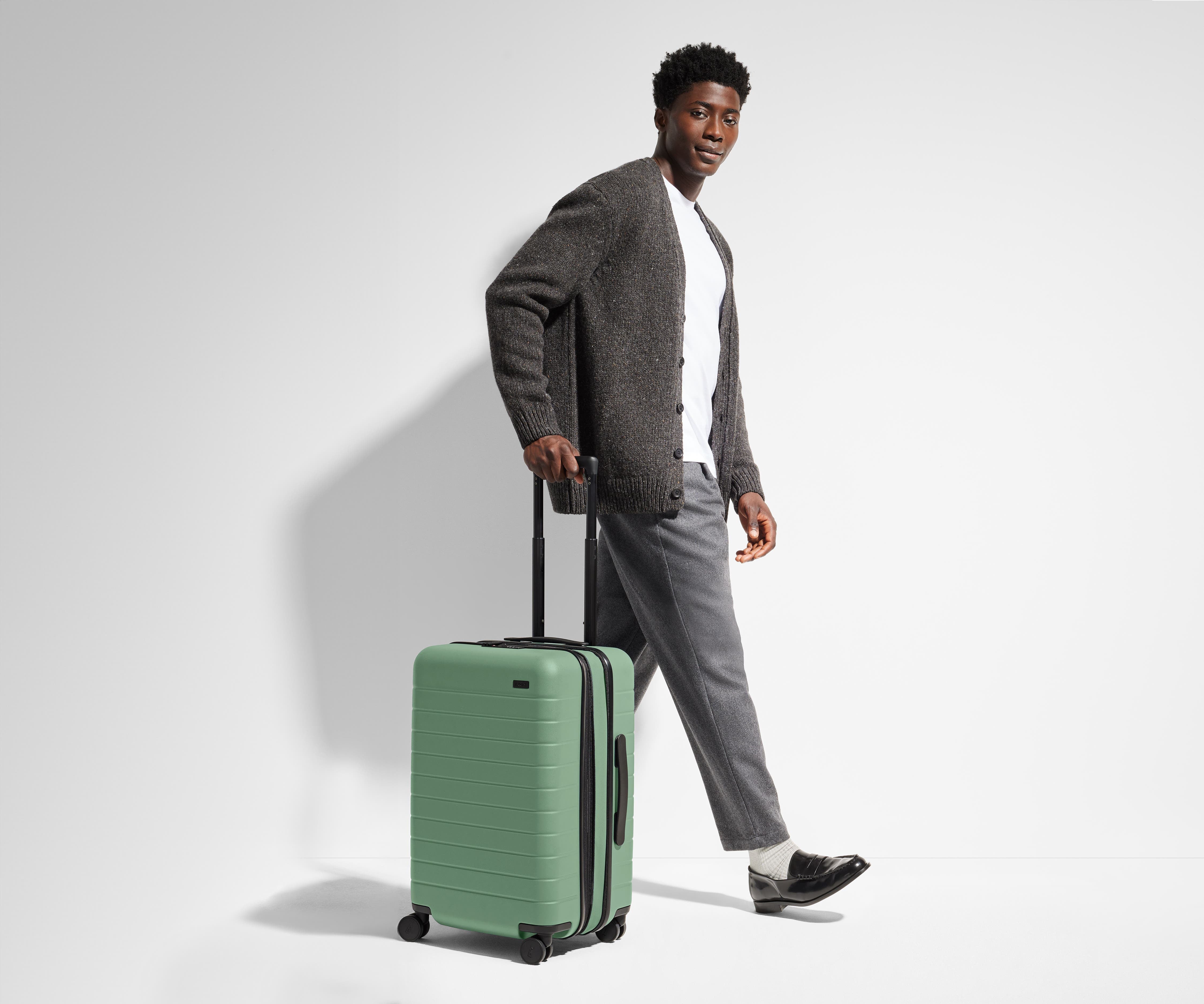 A 6'1" male model is standing next to and walking with The Bigger Carry-On suitcase by holding the raised handle to show size and scale