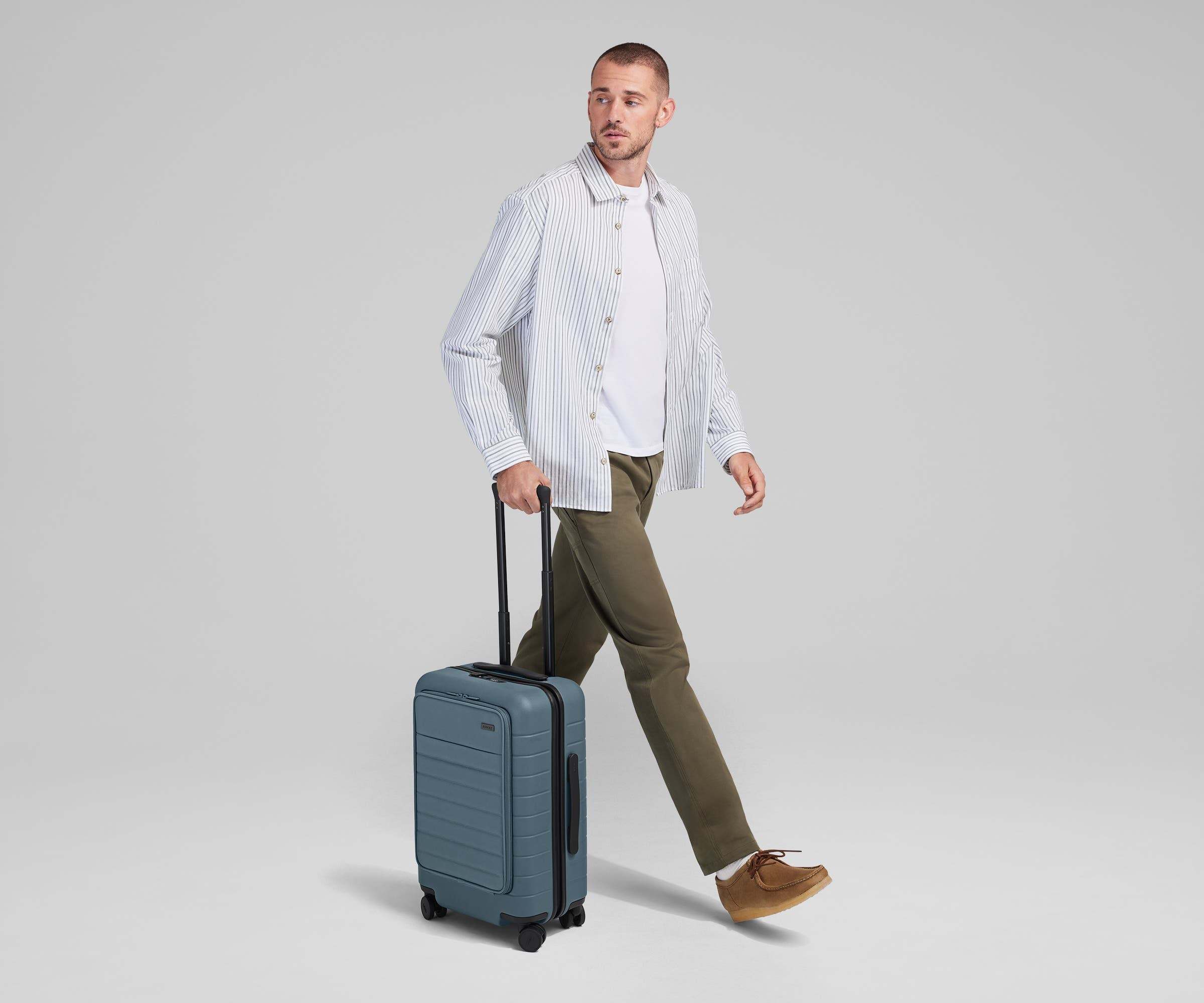 Male talent mid-stride, holding the trolley handle of The Front Pocket Carry-On in Coast Blue
