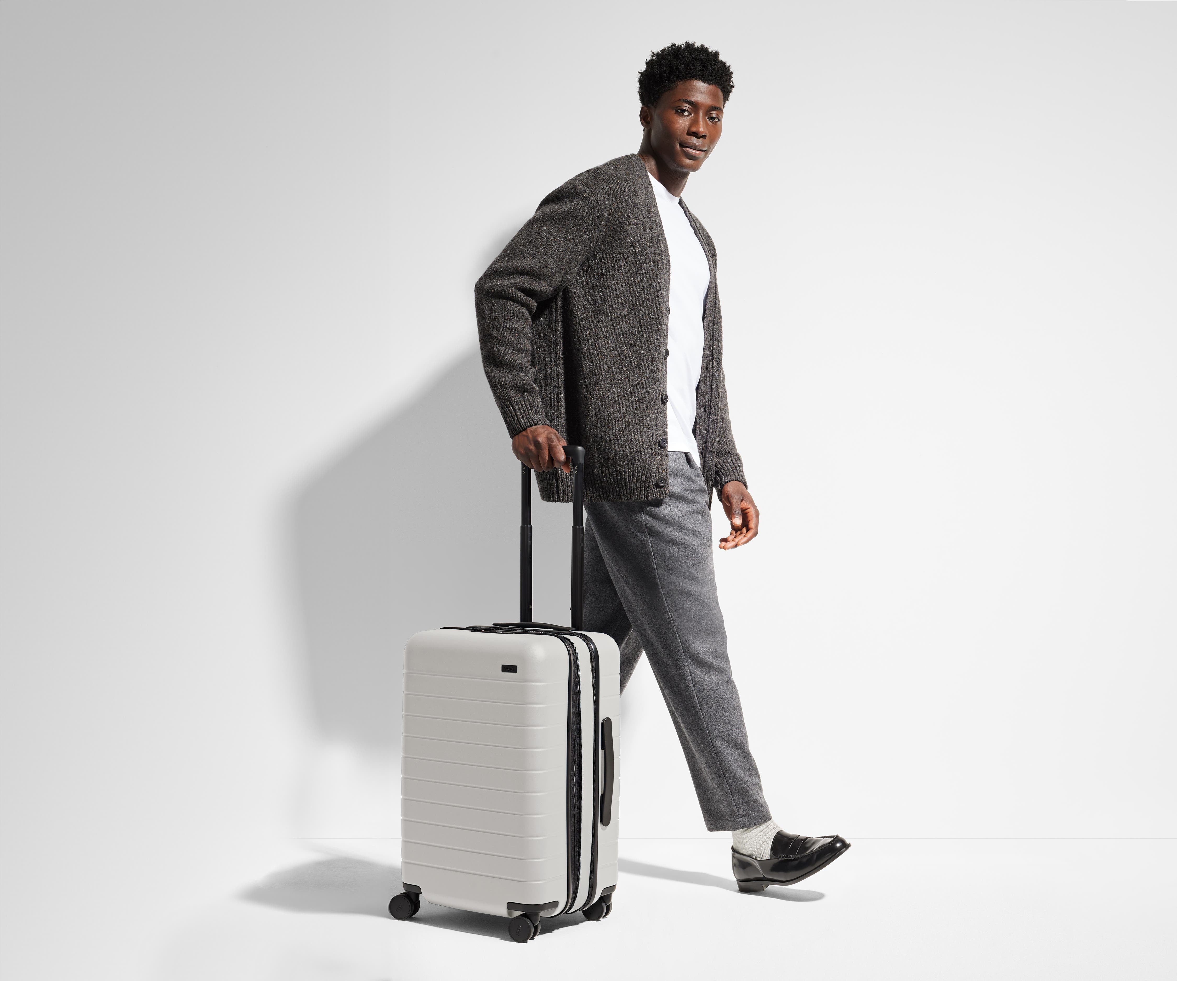 A 6'1" male model is standing next to and walking with The Bigger Carry-On suitcase by holding the raised handle to show size and scale