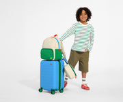 A child model holding the raised handle of The Kids Carry-On suitcase in Splash Blue with the Kids Backpack and Kids Lunch Box in Marshmallow White attached on top, completing the travel uniform