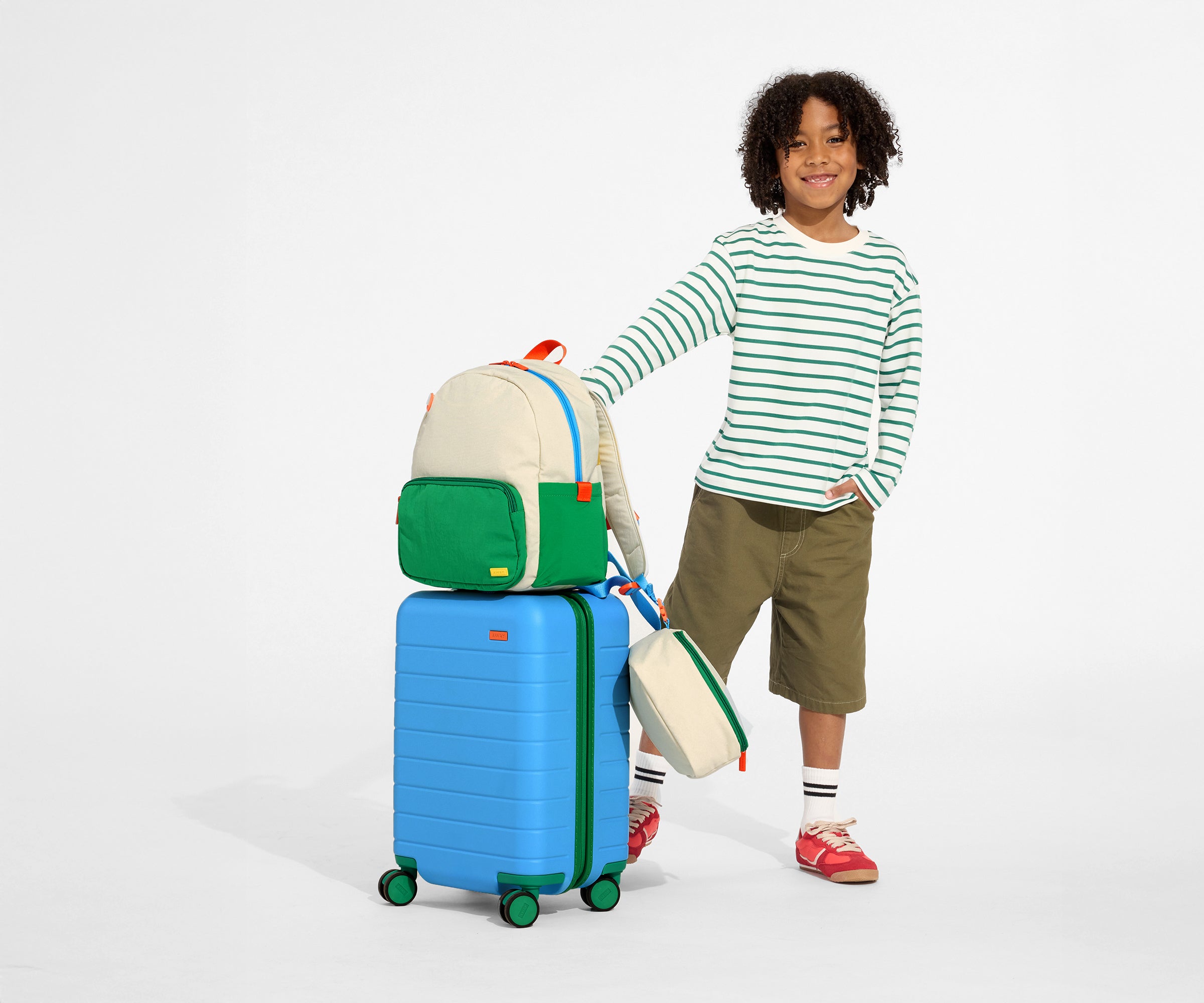 A child model holding the raised handle of The Kids Carry-On suitcase in Splash Blue with the Kids Backpack and Kids Lunch Box in Marshmallow White attached on top, completing the travel uniform