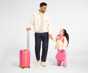 A child model holds hands with an adult model, who is pushing the raised handle of The Kids Carry-On suitcase in Cotton Candy Pink which is positioned at adult height, while holding the Kids Lunch Box in Cotton Candy Pink and wearing the Kids Backpack in Cotton Candy Pink