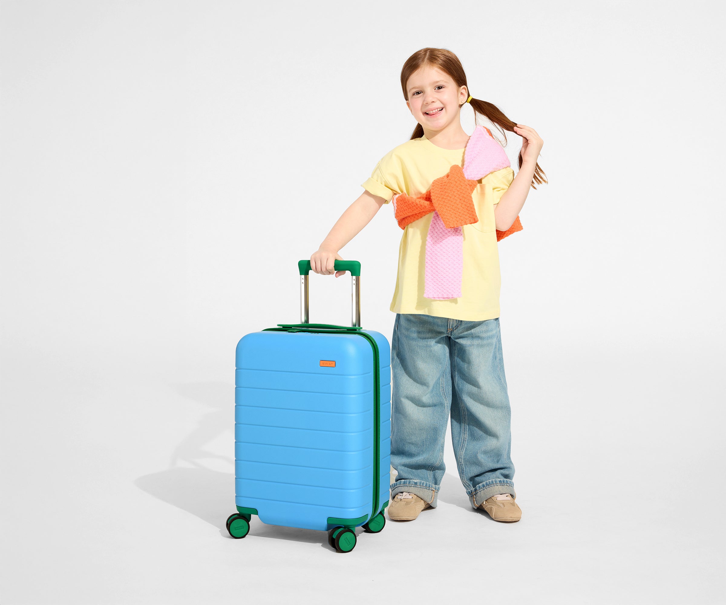 A child model is standing next to The Kids Carry-On suitcase in Splash Blue and holding onto the raised handle to show size and scale