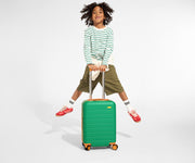 A child model holding the raised handle of The Kids Carry-On suitcase in Froggy Green and jumping in the air with a smile on their face