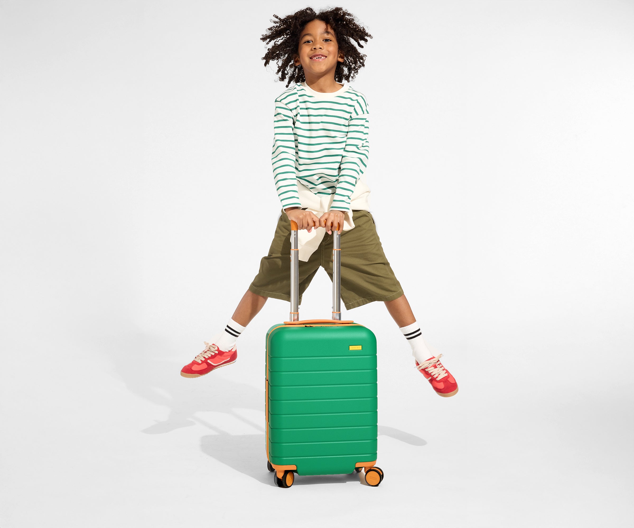 A child model holding the raised handle of The Kids Carry-On suitcase in Froggy Green and jumping in the air with a smile on their face