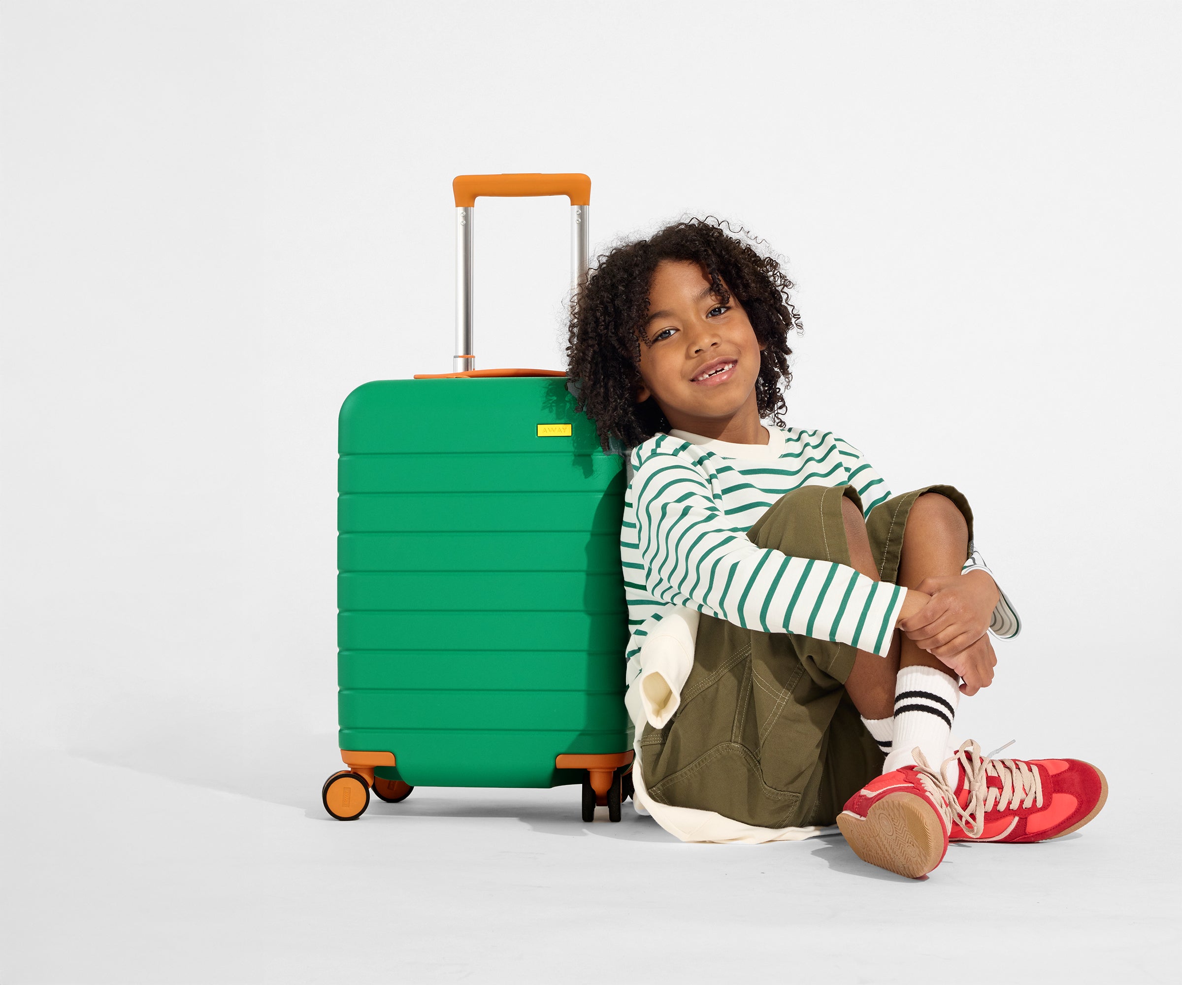 A child model sits on the floor and leans against The Kids Carry-On suitcase in Froggy Green