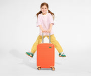 A child model holding the raised handle of The Kids Carry-On suitcase in Poppy Orange and jumping in the air with a smile on their face