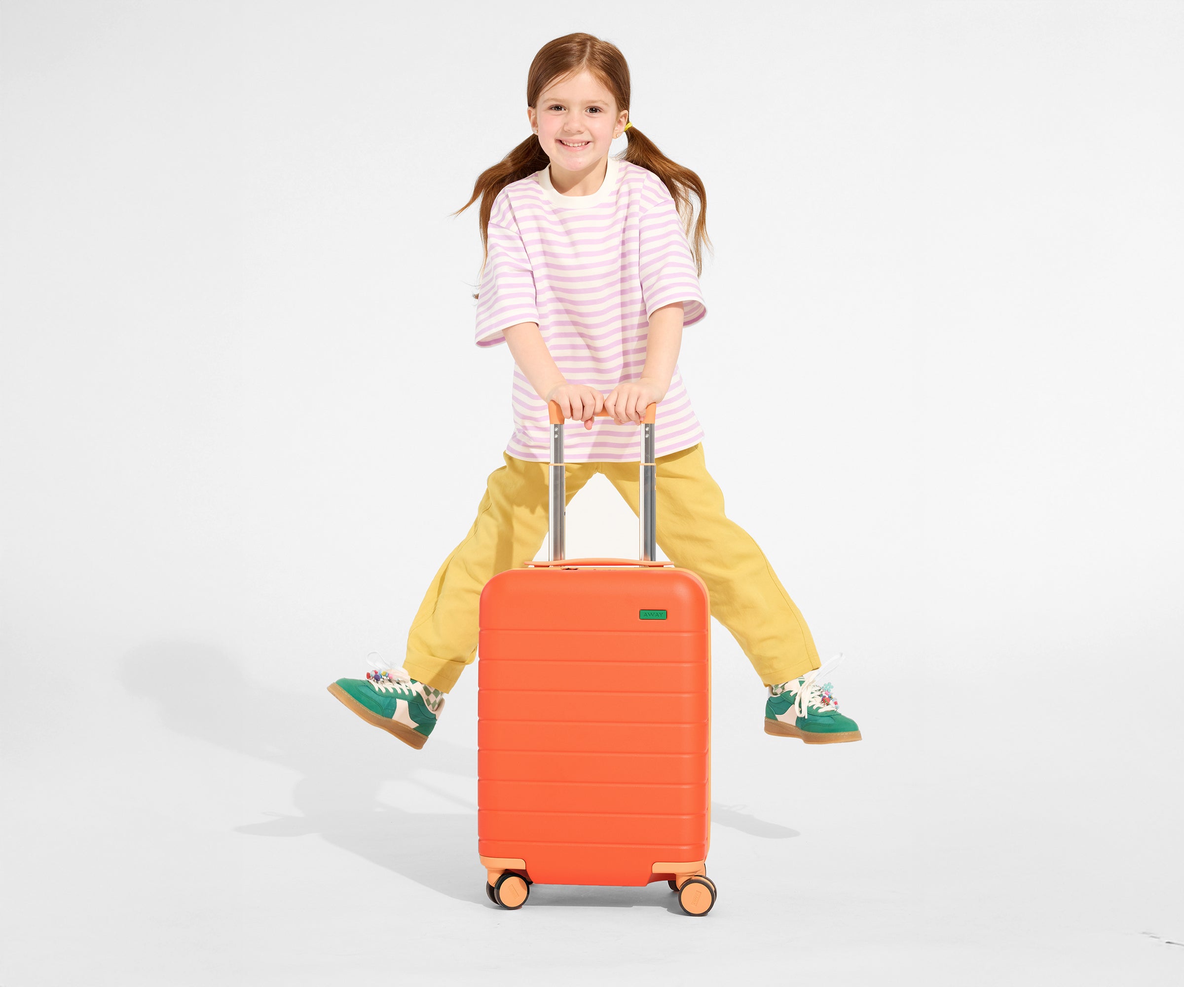 A child model holding the raised handle of The Kids Carry-On suitcase in Poppy Orange and jumping in the air with a smile on their face