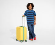A child model is standing next to The Kids Carry-On suitcase in Sunshine Yellow and holding onto the raised handle to show size and scale