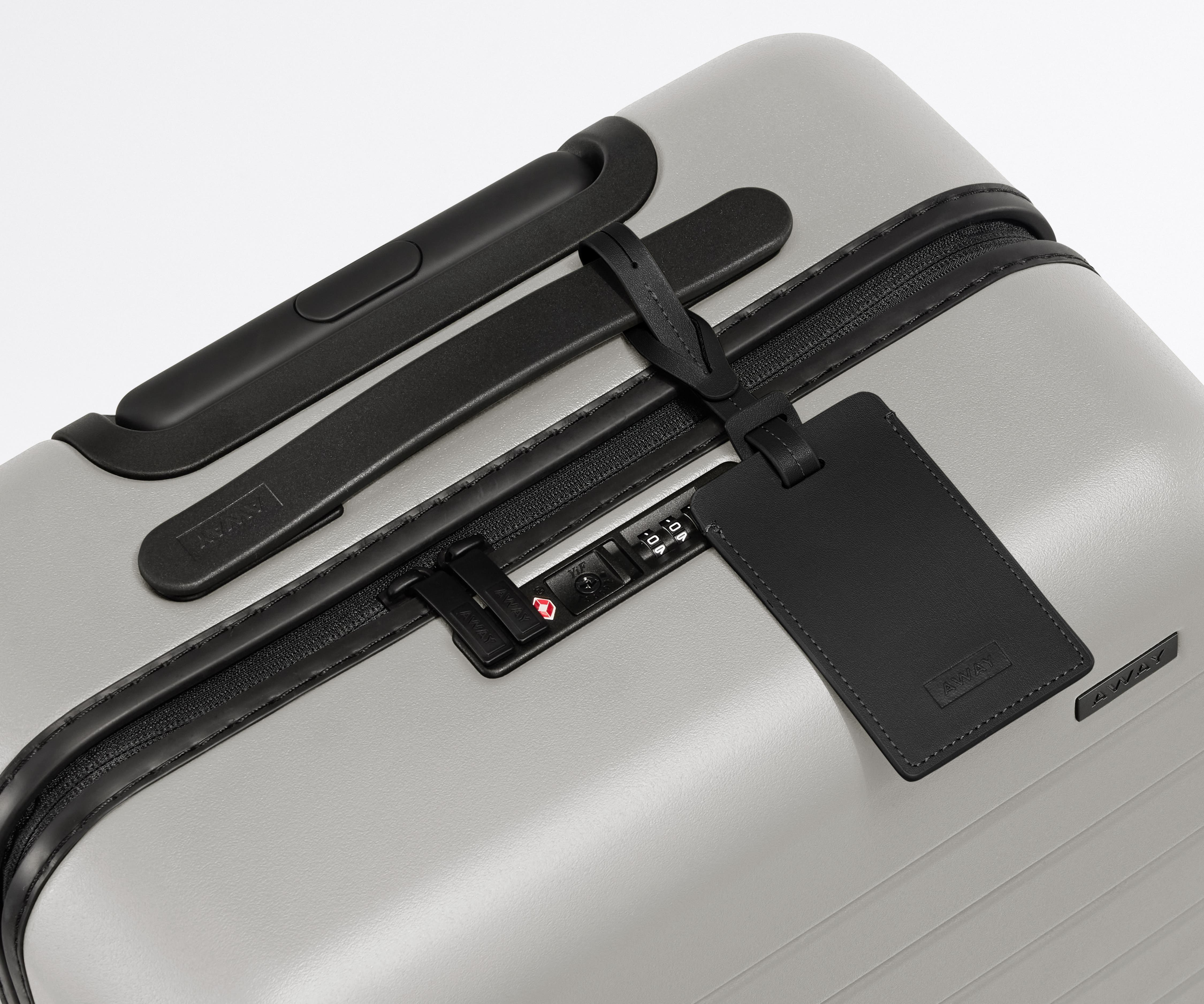 The Carry-On suitcase in Cloud Gray shown shown from above highlighting TSA-approved combination lock and recycled leather luggage tag