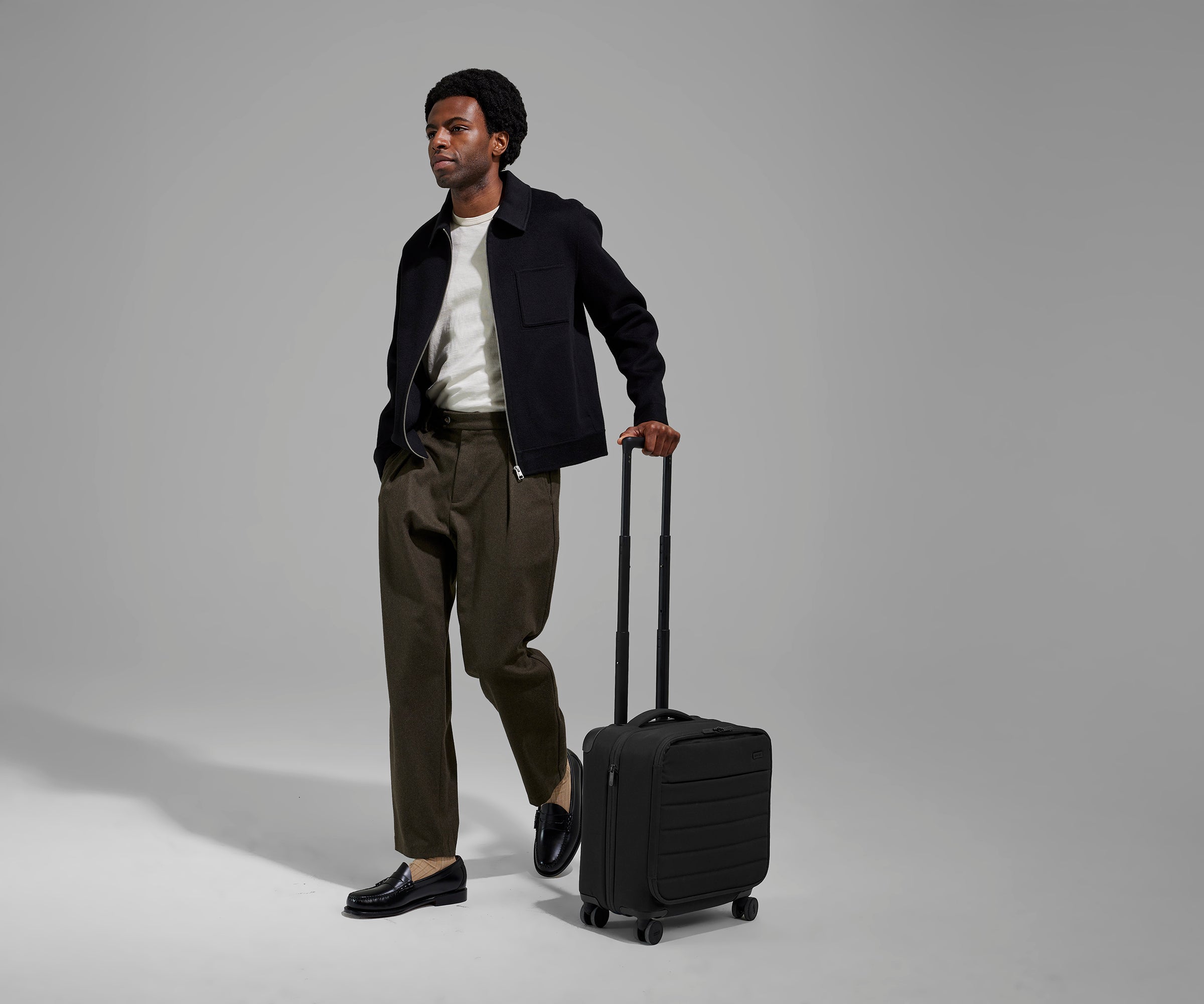 A 6'2" male model is standing next to The Softside Compact Carry-On suitcase in Jet Black and holding on to the raised handle to show size and scale