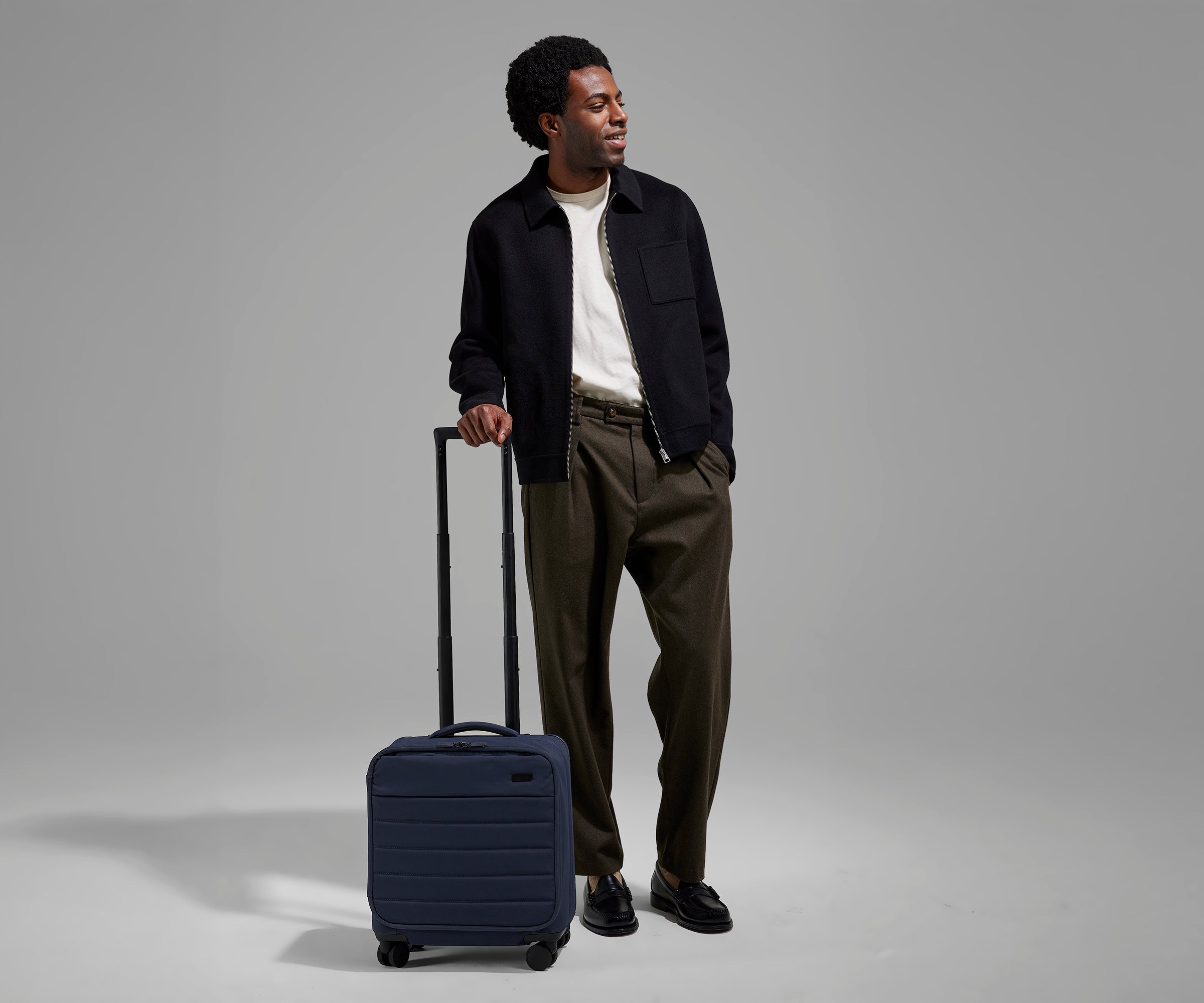 A 6'2" male model is standing next to The Softside Compact Carry-On suitcase in Navy Blue and holding on to the raised handle to show size and scale