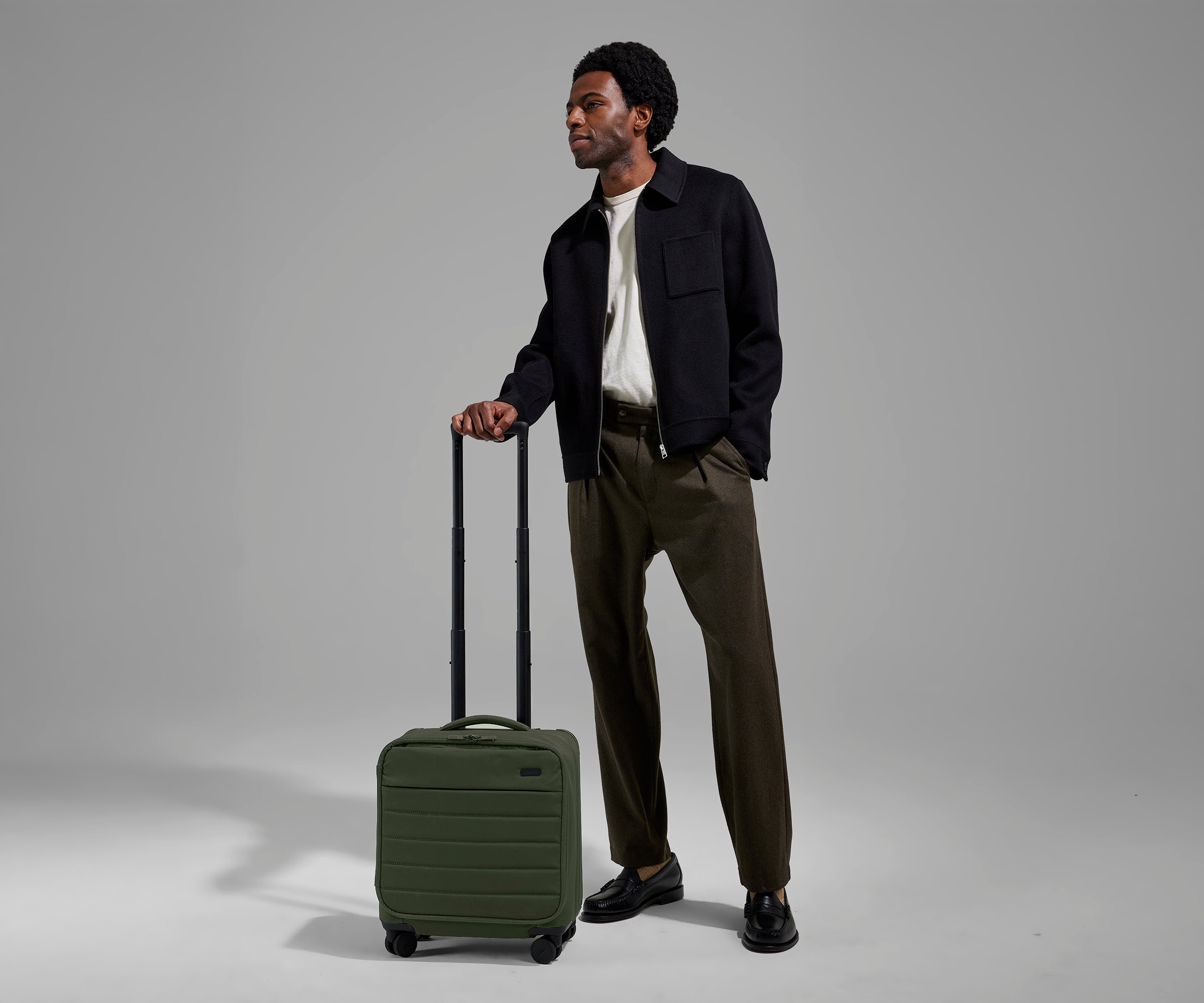 A 6'2" male model is standing next to The Softside Compact Carry-On suitcase in Olive Green and holding on to the raised handle to show size and scale