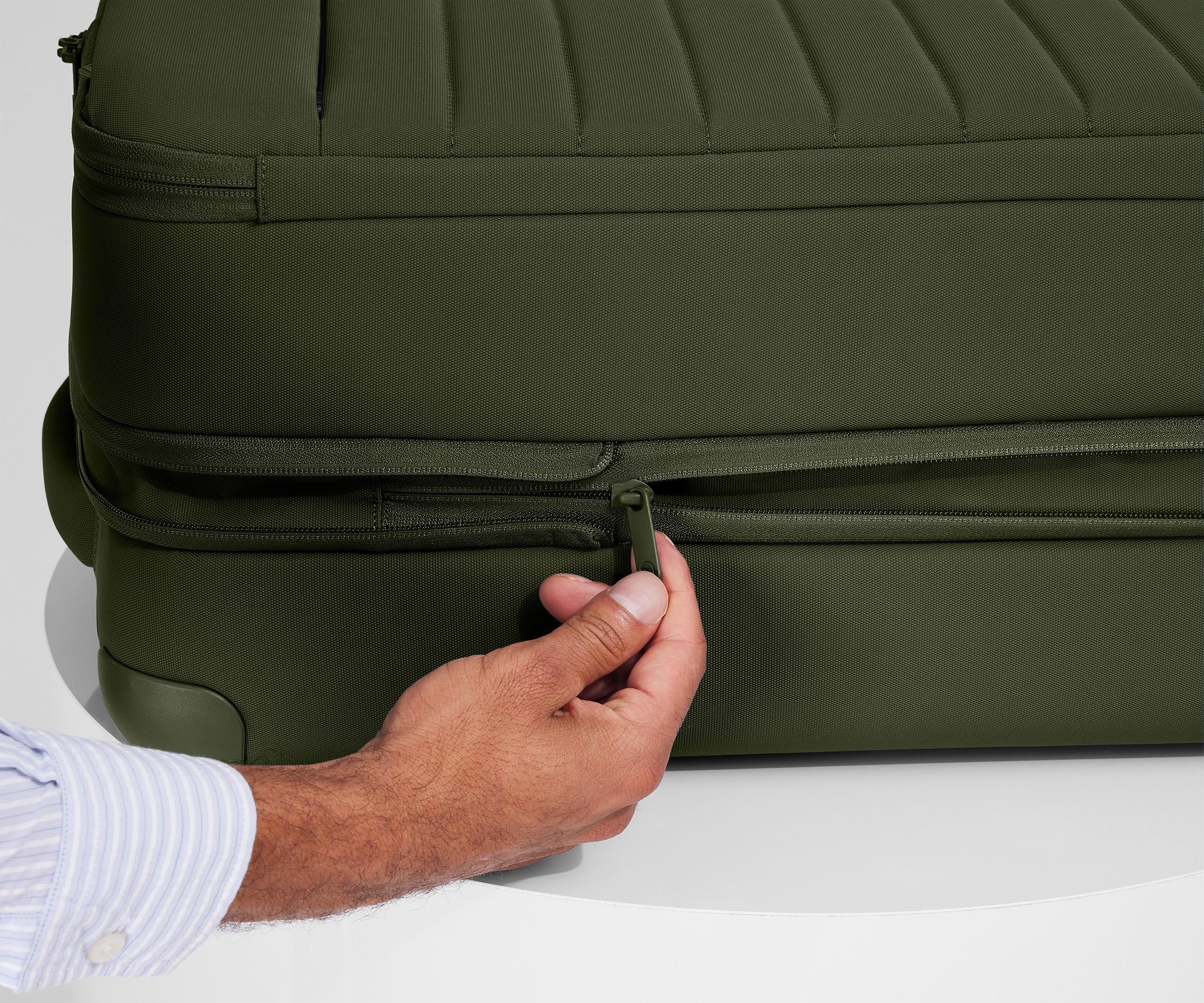 A person is unzipping The Softside Bigger Carry-On suitcase in Olive Green using the wraparound zipper to access 1.75" (4.5 cm) of extra packing space