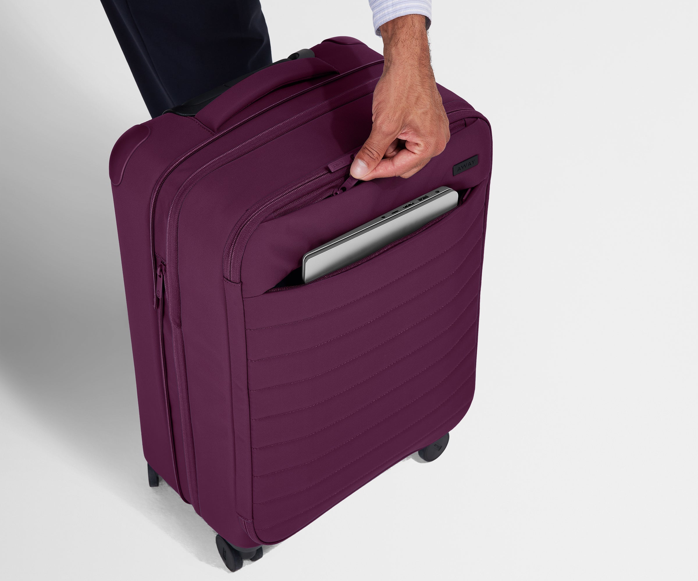 A person is unzipping the hidden exterior pocket on The Softside Carry-On in Plum Purple, and there is a laptop peeking out of the second, deeper no-show pocket on the outside of the suitcase.