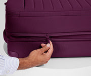A person is unzipping The Softside Bigger Carry-On suitcase in Plum Purple using the wraparound zipper to access 1.75" (4.5 cm) of extra packing space