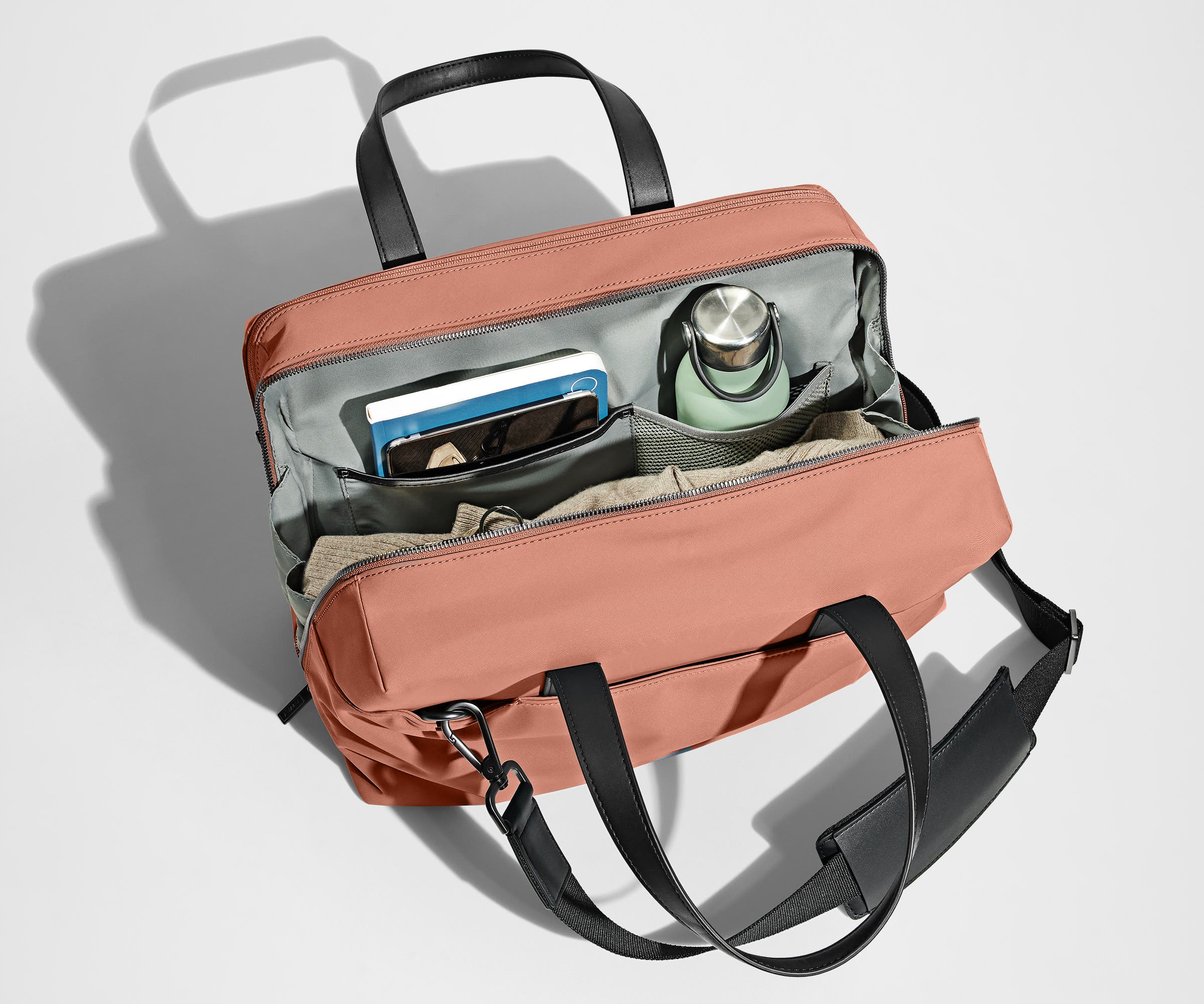 Main compartment packed with a light sweater, notebook, tablet, and water bottle. The detachable shoulder strap is clipped onto the bag.