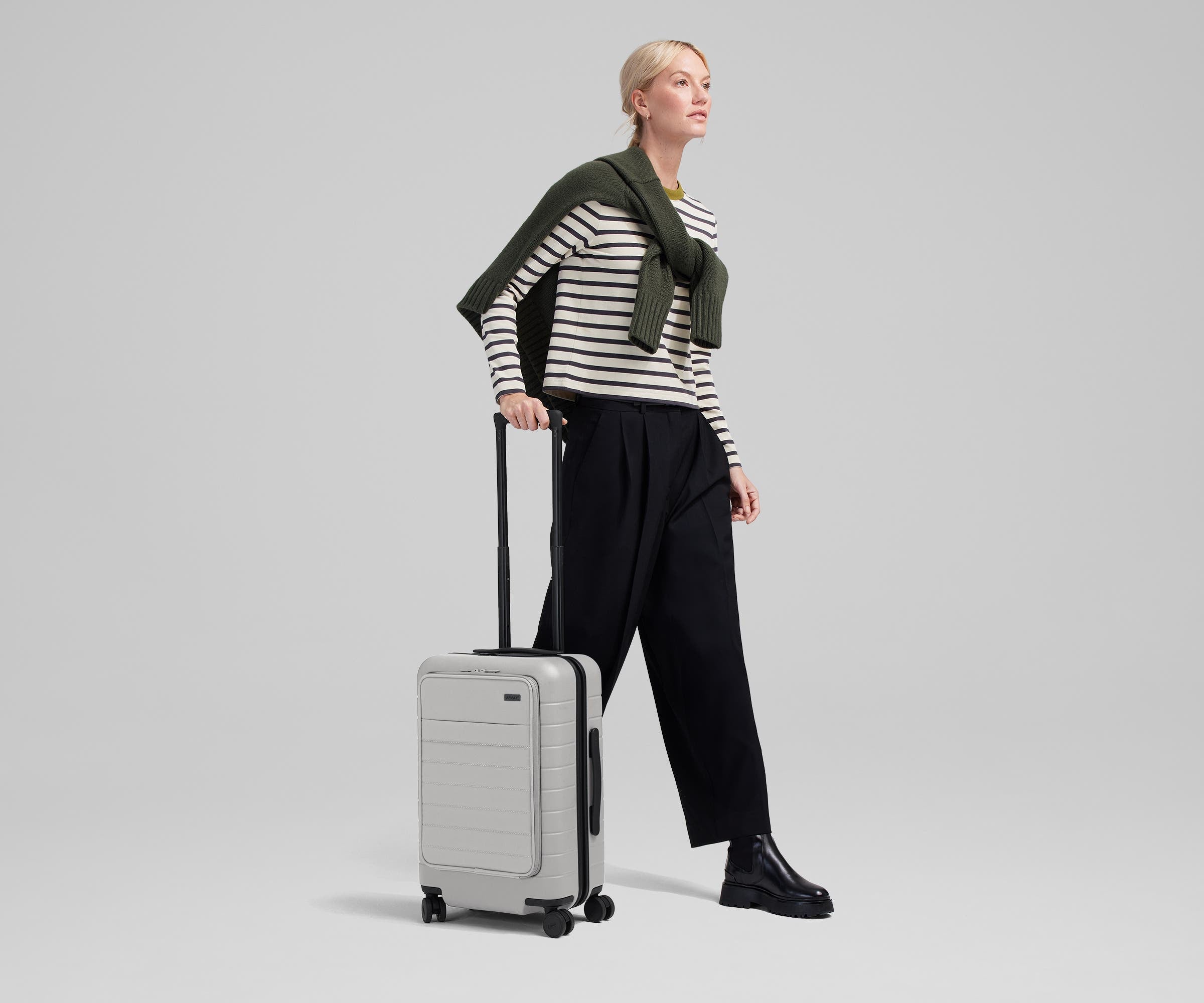 Female talent mid-stride, holding the trolley handle of The Front Pocket Carry-On in Cloud Gray