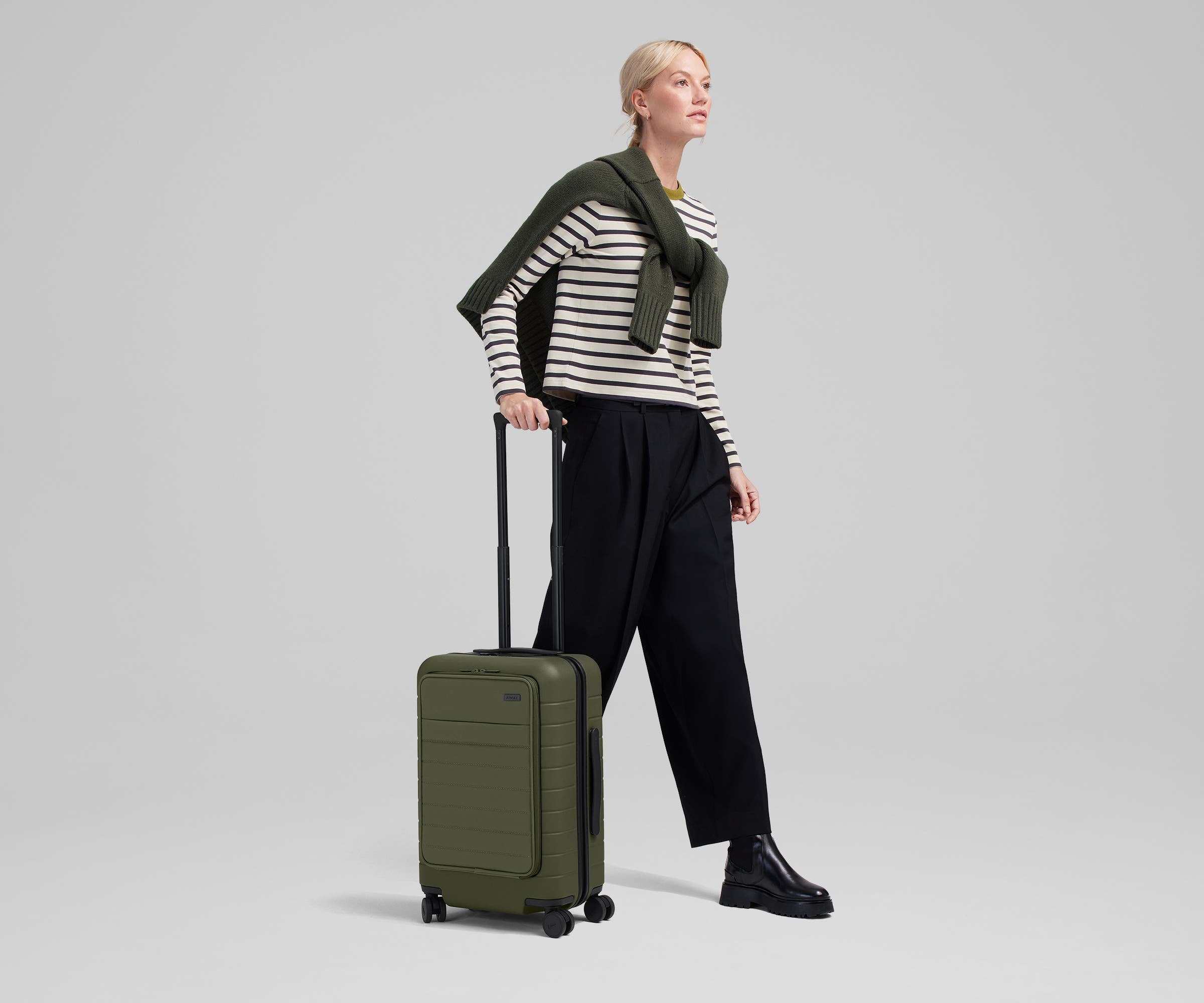 Female talent mid-stride, holding the trolley handle of The Front Pocket Carry-On in Olive Green