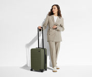 A 5'6" female model is standing next to The Carry-On suitcase and holding onto the raised handle to show size and scale