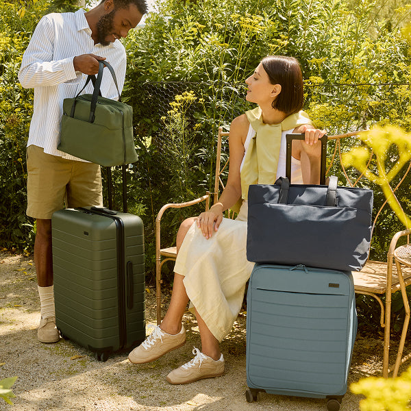 A man and woman sitting outside amongst trees with sets of Away luggage in green and blue