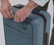 A video of a person packing the compartments of the front pocket and hidden access pocket with a laptop, tablet, and other essentials of The Front Pocket Bigger Carry-On suitcase in Coast Blue