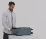 A 6'0" male model filling The Softside Bigger Carry-On suitcase in Coast Blue with Away's packing organizers and using the interior organization and compression system for seamless packing